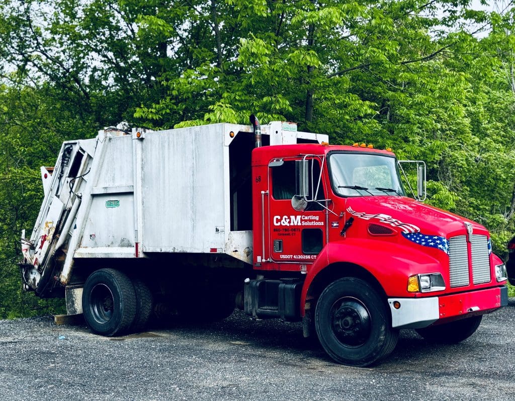 Red garbage truck with patriotic white and blue star decals, parked on a gravel lot with green trees in the background.