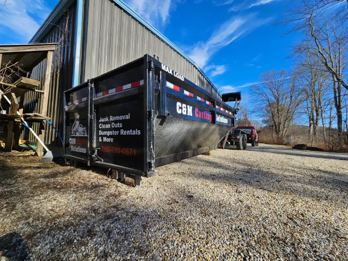 Large black dumpster trailer labeled "C&M Carting Solutions" with a pickup truck attached, parked on a gravel lot beside a metal building.